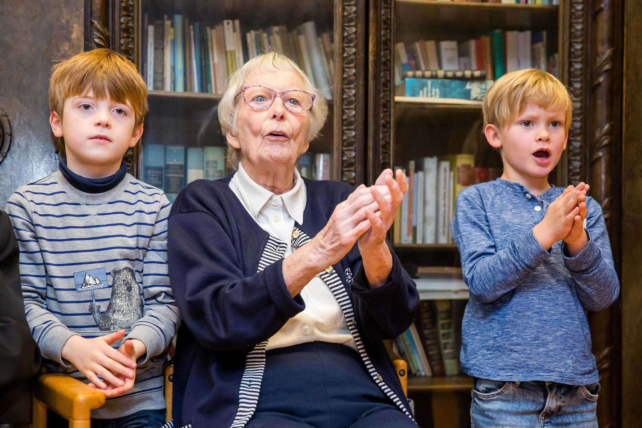 Zwei kleine Jungs klatschen zusammen mit einer älteren Dame. Alle drei blicken in die selbe Richtung. Im Hintergrund ist ein Bücherregal zu sehen
