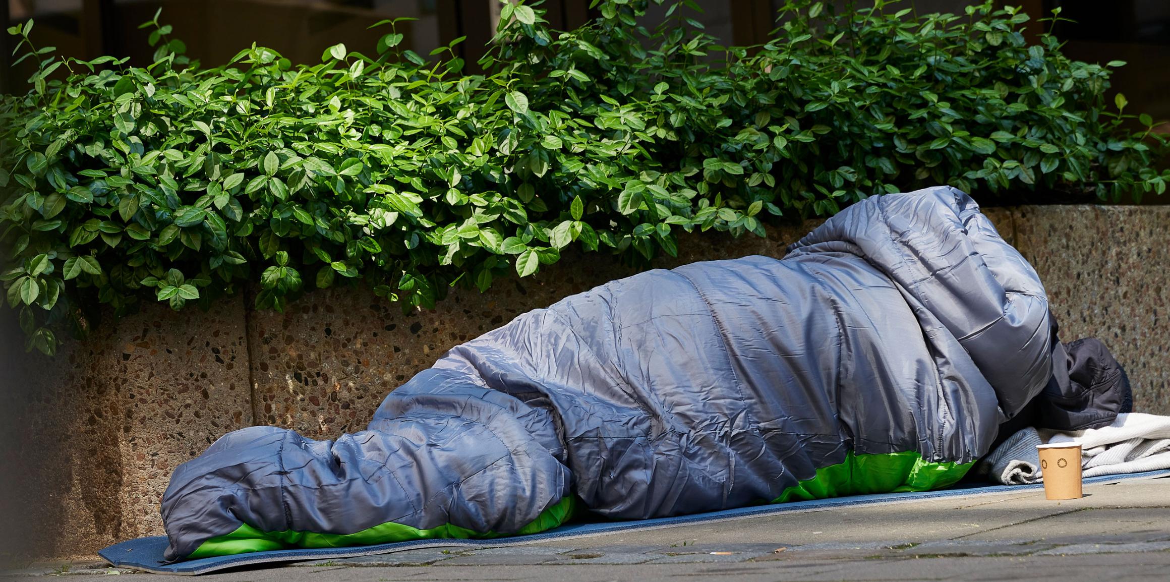 Mann liegt auf der Straße im Schlafsack. Er ist obdachlos. Vor ihm steht ein Becher. Wahrscheinlich, um nach Geld zu bitten.