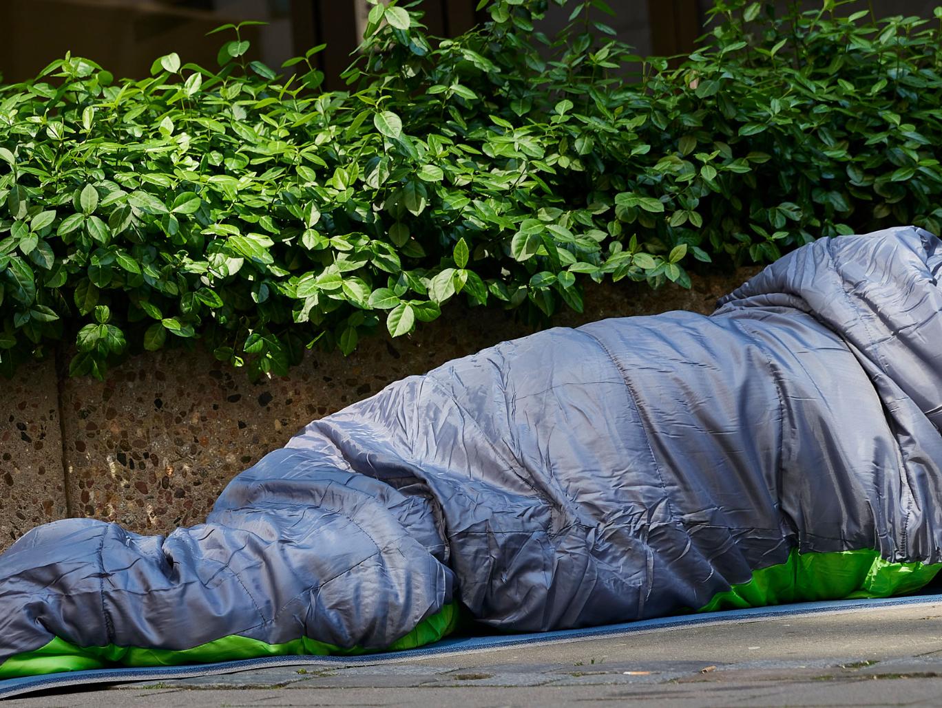 Mann liegt auf der Straße im Schlafsack. Er ist obdachlos. Vor ihm steht ein Becher. Wahrscheinlich, um nach Geld zu bitten.