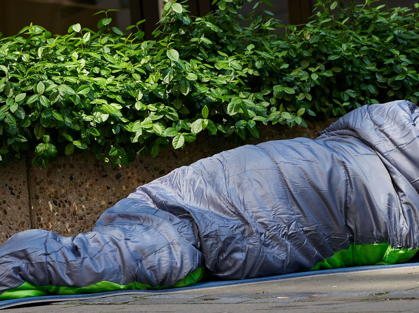Mann liegt auf der Straße im Schlafsack. Er ist obdachlos. Vor ihm steht ein Becher. Wahrscheinlich, um nach Geld zu bitten.