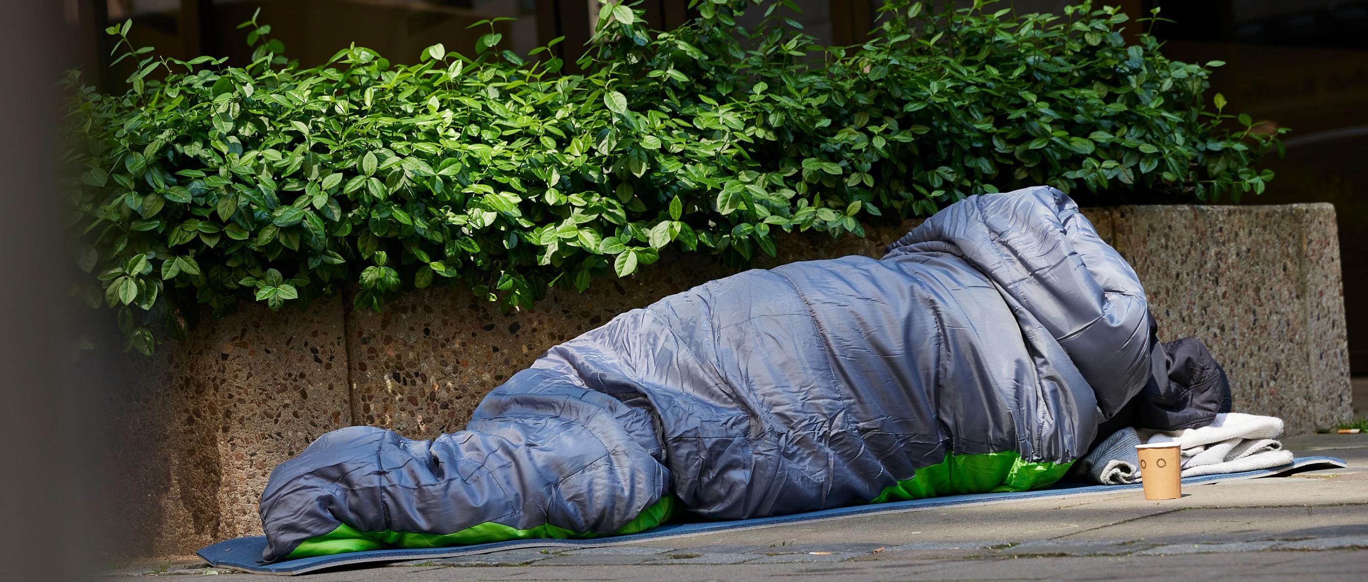 Mann liegt auf der Straße im Schlafsack. Er ist obdachlos. Vor ihm steht ein Becher. Wahrscheinlich, um nach Geld zu bitten.