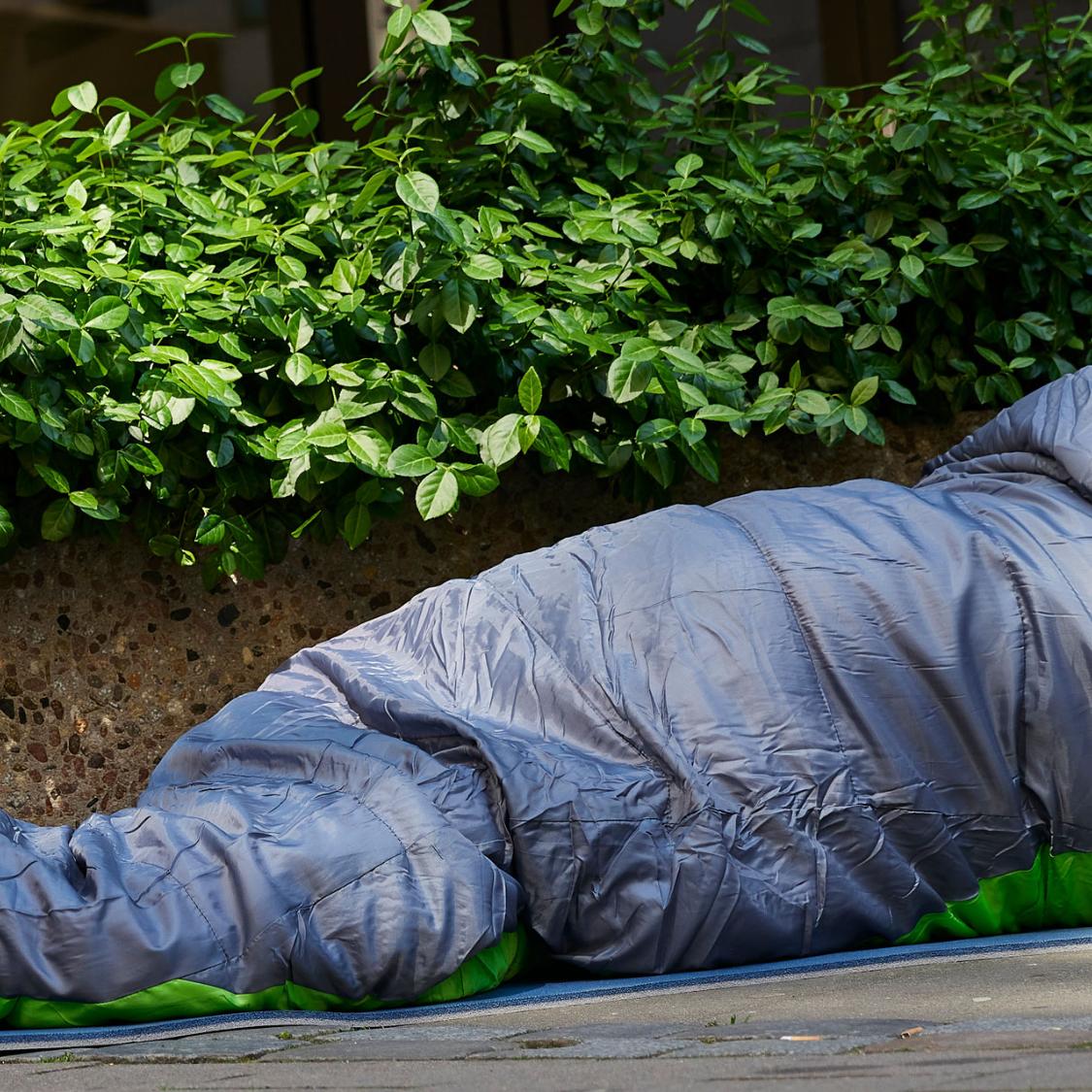 Mann liegt auf der Straße im Schlafsack. Er ist obdachlos. Vor ihm steht ein Becher. Wahrscheinlich, um nach Geld zu bitten.