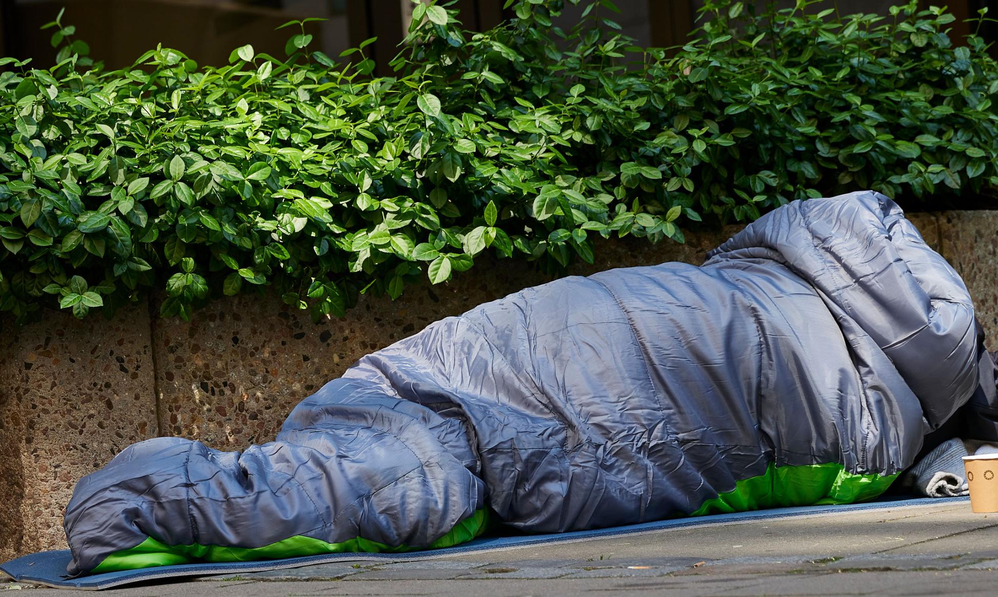 Mann liegt auf der Straße im Schlafsack. Er ist obdachlos. Vor ihm steht ein Becher. Wahrscheinlich, um nach Geld zu bitten.